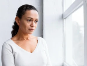 Mujer de mediana edad mirando por la ventana con expresión de preocupación y estrés por falta de empleo y síntomas de depresión.