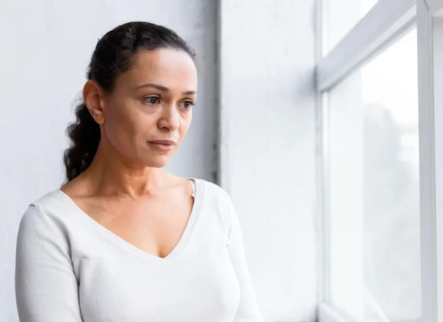 Mujer de mediana edad mirando por la ventana con expresión de preocupación y estrés por falta de empleo y síntomas de depresión.