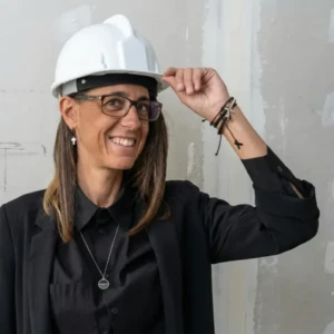 Retrato de la arquitecta Lucía Toro sonriendo, vistiendo un casco de seguridad blanco y saco negro frente a un muro en construcción.